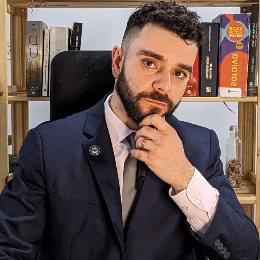 Israel M. Mesquita, advogado com terno azul e gravata prateada, sentado em seu escritório com uma prateleira de livros ao fundo, com a mão no queixo em uma pose que transmite profissionalismo.